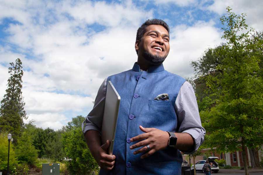 person holding a laptop wearing a blue vest and shirt standing in front of fir trees